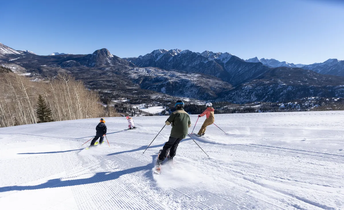 A dynamic exterior shot featuring skiers gliding down pristine, snow-covered slopes with the mountains and a bright blue sky as a backdrop. The lively scene illustrates the thrill and joy of slope access at Purgatory, emphasizing the excitement and beauty of the winter landscape.