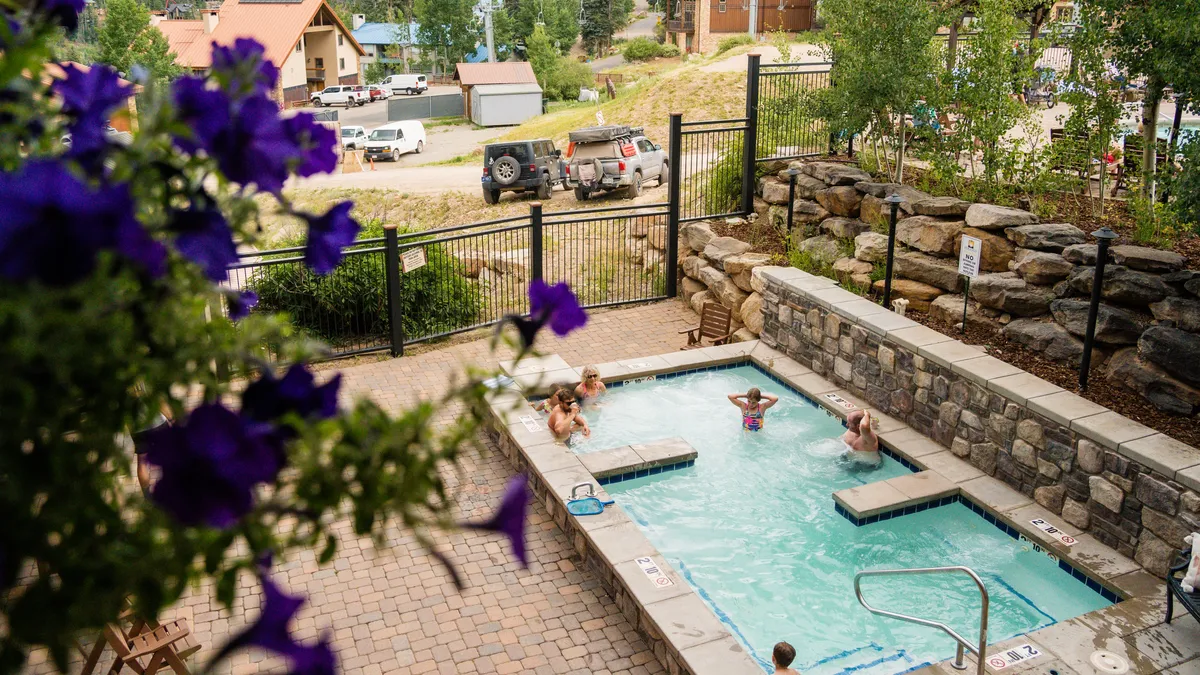 A vibrant outdoor swimming pool surrounded by lush greenery, with people lounging and swimming. The pool area is lively, evoking a sense of fun and relaxation, perfect for families and guests looking to unwind after a day on the slopes.