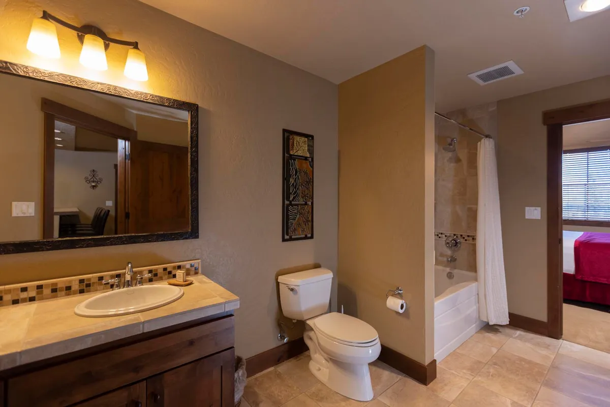 A well-appointed bathroom featuring a vanity with a sink and framed mirror, decorative wall art, and a bathtub with an overhead shower. Warm lighting and tasteful decor create a tranquil, inviting mood, ideal for relaxation after a day of mountain adventures.