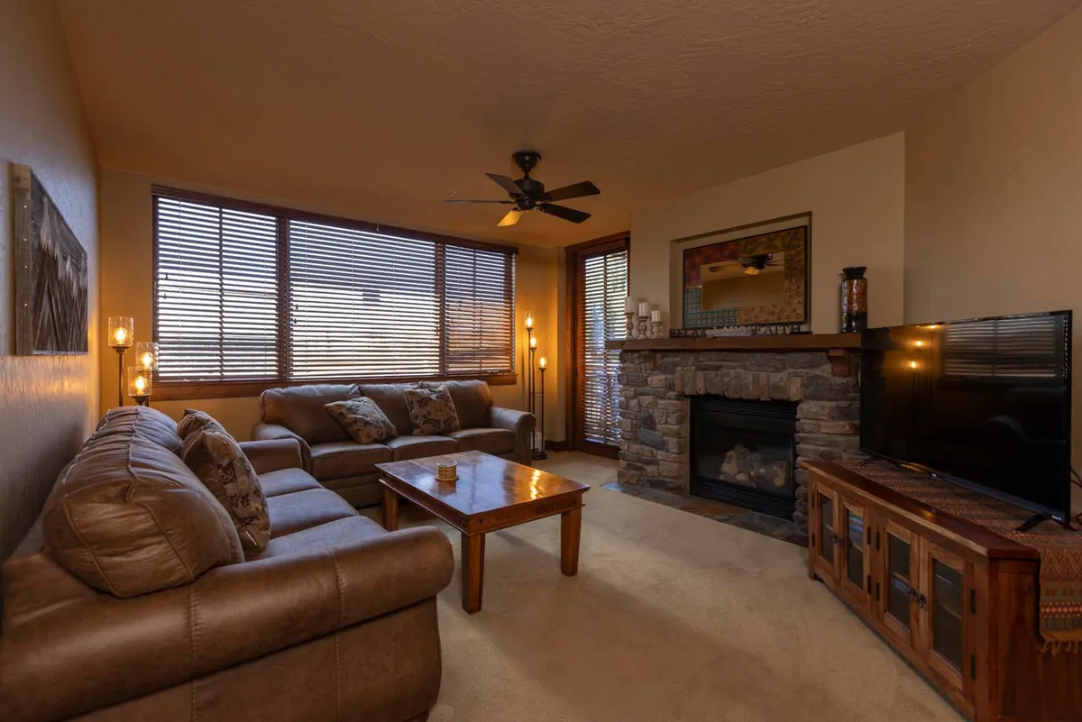 The hero image features a cozy living area with a striking stone fireplace, plush and inviting seating, and expansive windows that frame breathtaking mountain views. The warm lighting and natural materials create a welcoming atmosphere perfect for gathering with friends or family after a day on the slopes.