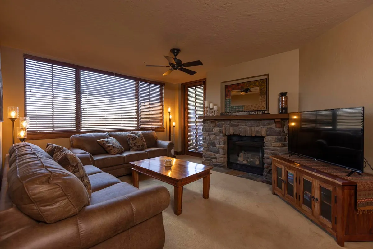 A spacious living room with comfortable leather sofas arranged around a rustic stone fireplace. Large windows with blinds allow natural light to flood the space, and a wooden coffee table sits at the center. A television is mounted for entertainment.