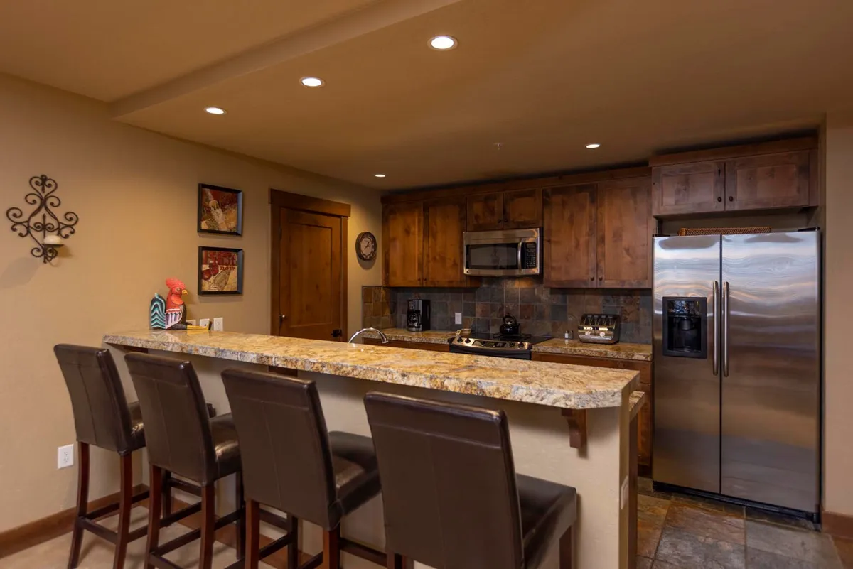 A cozy, well-lit kitchen featuring a granite countertop with bar stools, modern kitchen cabinets, and stainless steel appliances. Decorative wall art adds warmth, while the open layout encourages both cooking and casual dining or conversation.