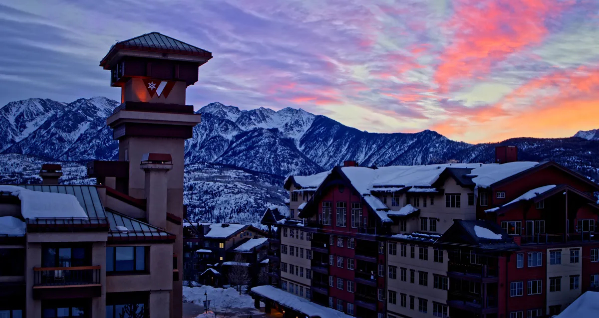 An exterior view of Purgatory Lodge framed by the dramatic, snow-covered Needles Mountain Range under a colorful sky. The village buildings and iconic clock tower evoke a vibrant mountain resort atmosphere, perfect for year-round adventure.