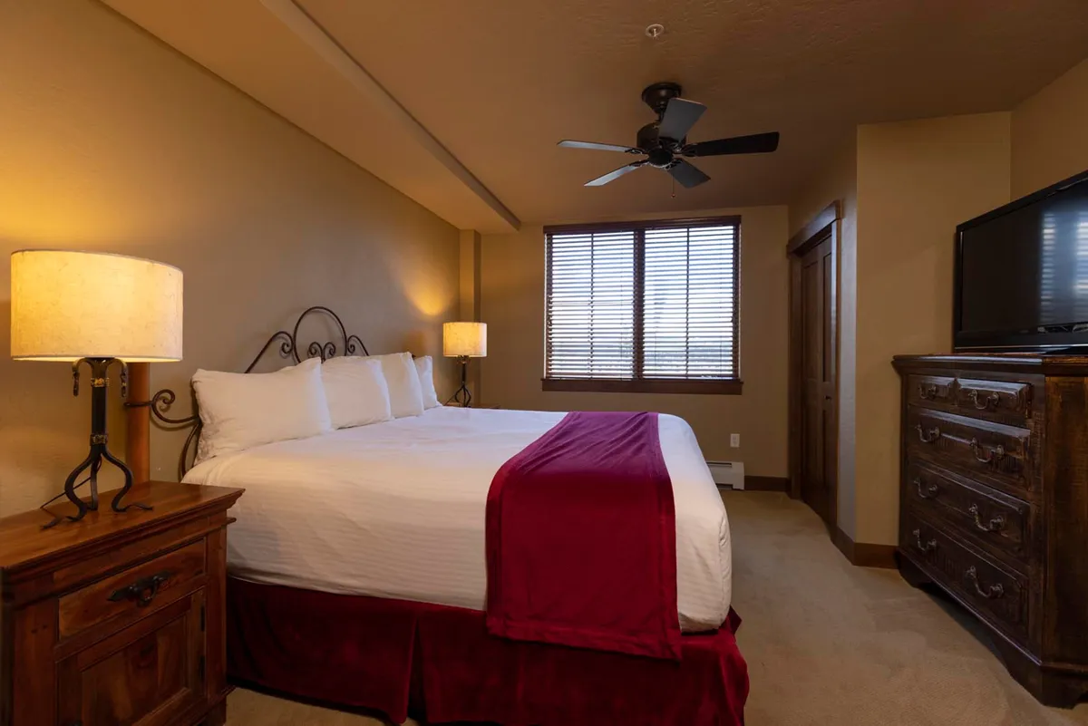 A comfortable bedroom showcasing a bed dressed with a red throw, a wooden nightstand and lamp, and a window with partially open blinds. A wooden dresser is also visible, creating a sense of homey comfort and warmth, ideal for unwinding after a day outdoors.