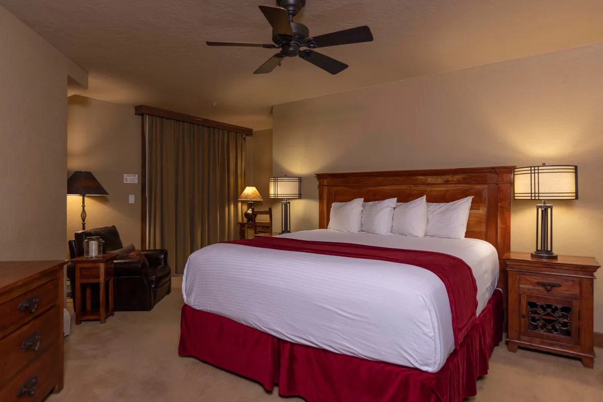 A warmly lit bedroom featuring a king-sized bed with crisp linens, flanked by matching nightstands and lamps. The soft glow creates an inviting atmosphere, emphasizing comfort and relaxation. A dresser sits at the foot of the bed, reinforcing the room's practical yet cozy design.