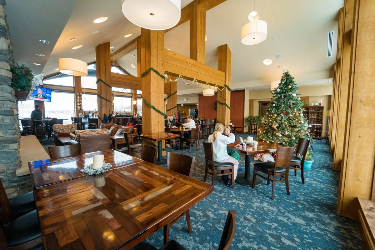 A festive dining area with a large table set for a family gathering, warm lighting, a decorated Christmas tree, and a bar area in the background. Large windows let in natural light, enhancing the inviting ambiance.