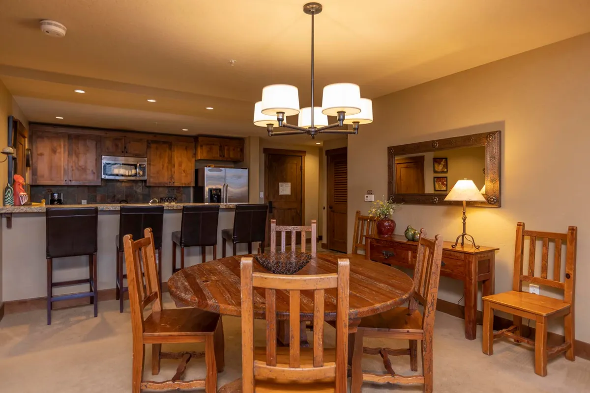 An inviting dining area featuring a wooden table set for a meal, adjacent to the kitchen. A chandelier hangs overhead, and a mirror with a lamp adds warmth and depth to the space.